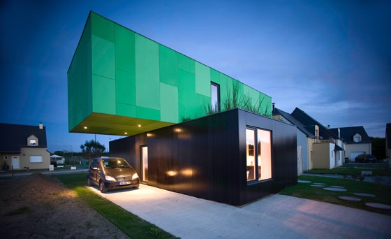 Modern two-story house with green upper section and car parked in driveway during evening. Unique contemporary architecture.