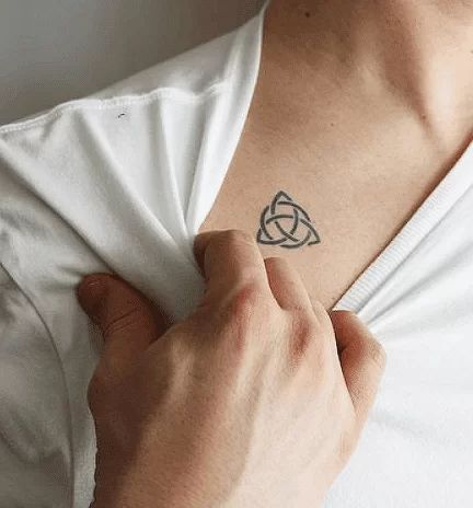 Person revealing a Celtic knot tattoo on their chest by pulling down a white shirt.