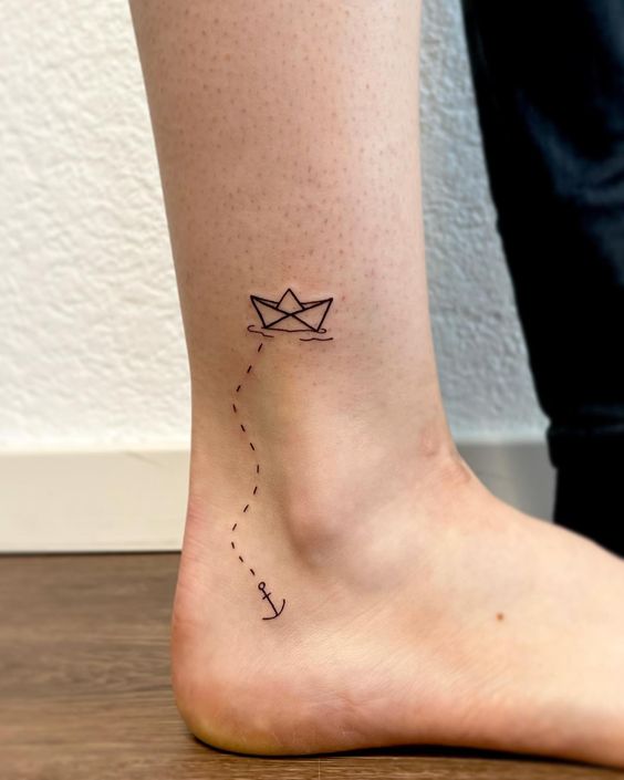 Minimalist paper boat and anchor tattoo on the ankle, featuring a dotted path connecting the two symbols.