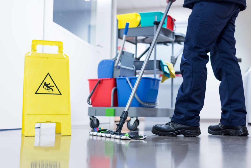 Janitor mopping shiny floor near wet floor sign and cleaning cart in office or commercial building.