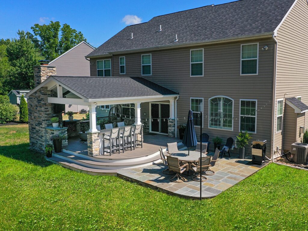 Backyard with covered patio, outdoor kitchen, stone fireplace, dining area, and lush green lawn on a sunny day.