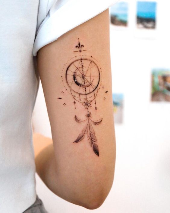 Elegant dreamcatcher tattoo with detailed feathers and crescent moon design on a person's upper arm. Minimalistic and artistic.