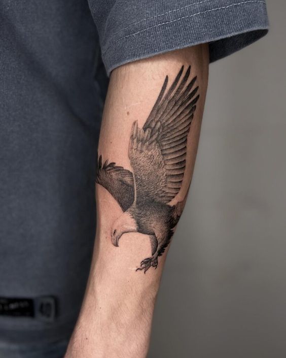 Detailed black and grey eagle tattoo on forearm, soaring with wings outstretched. Realistic design, impressive arm ink.