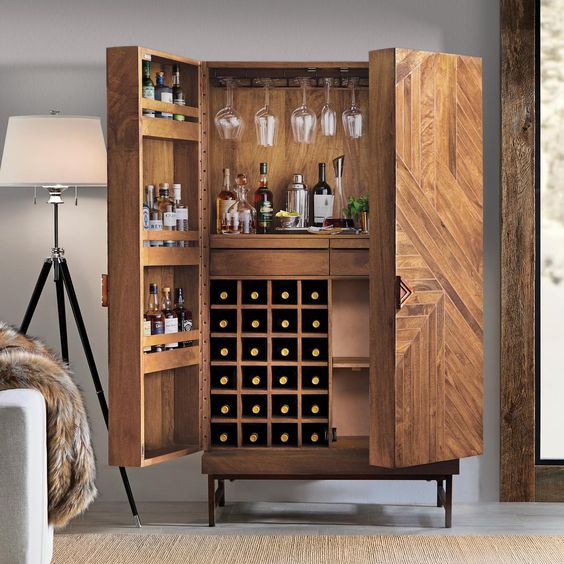 Open wooden bar cabinet with wine storage, liquor bottles, hanging glassware, and cocktail accessories, beside a floor lamp.