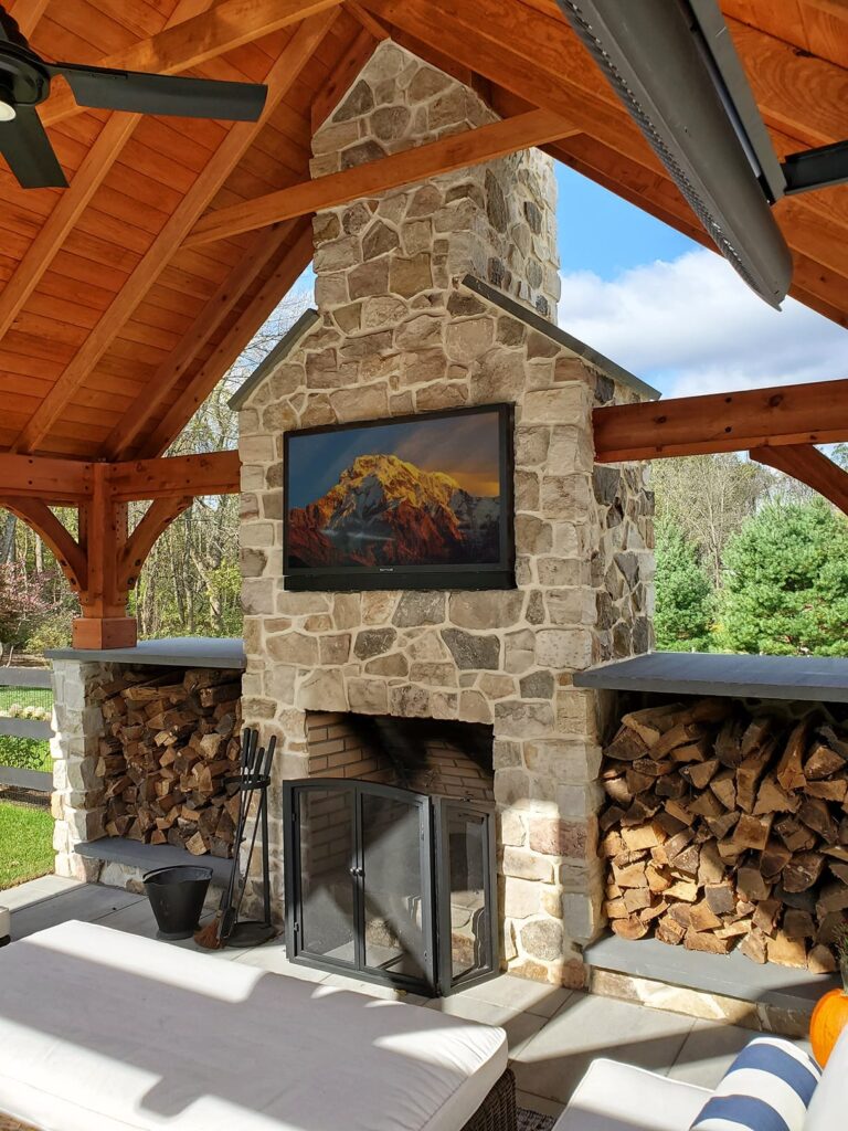 Outdoor stone fireplace with firewood storage under a wooden pergola, featuring a TV screen displaying a mountain landscape.