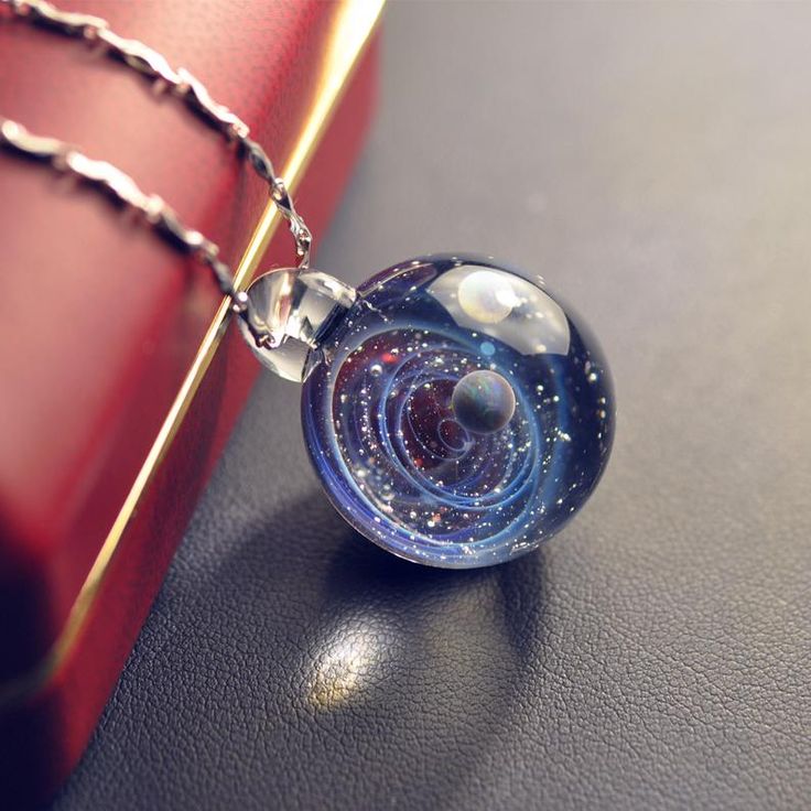 Close-up of a glass orb necklace featuring a miniature galaxy design, placed next to a red gift box on a dark surface.