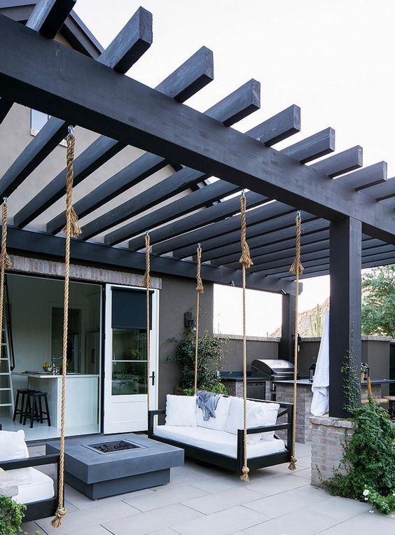 Modern patio with black pergola, hanging sofas, sleek grey fire pit, and outdoor kitchen area for stylish backyard living.