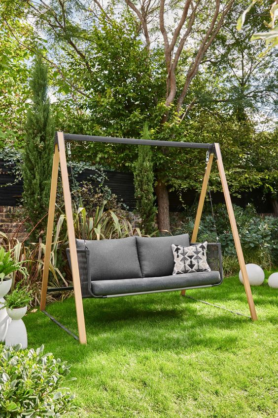 Outdoor garden swing with grey cushions and a decorative pillow, surrounded by lush greenery and vibrant plants.