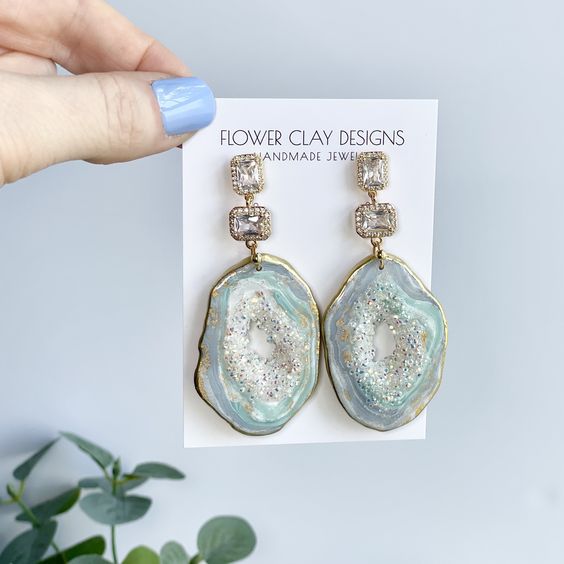 Hand holding handmade geode earrings from Flower Clay Designs, featuring glittering stones and blue accents.