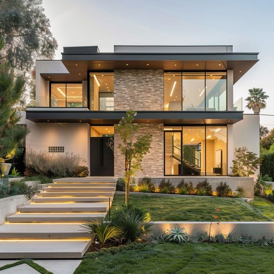 Modern two-story home with large glass windows, landscaped garden, and illuminated front steps at dusk.