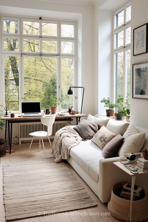 Bright home office with large windows, cozy white sofa, houseplants, and a desk with a laptop, creating a serene workspace.