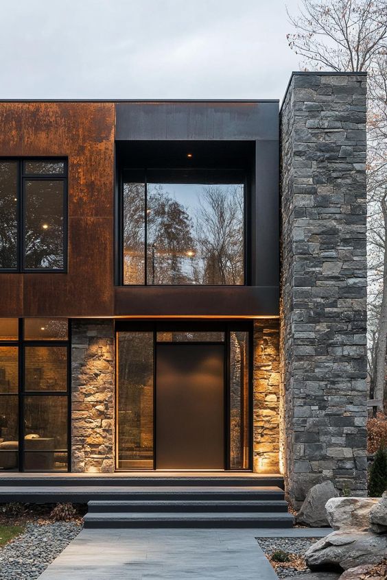 Modern house exterior with stone and rusted metal facade, large windows, stylish lighting, and a sleek black front door.