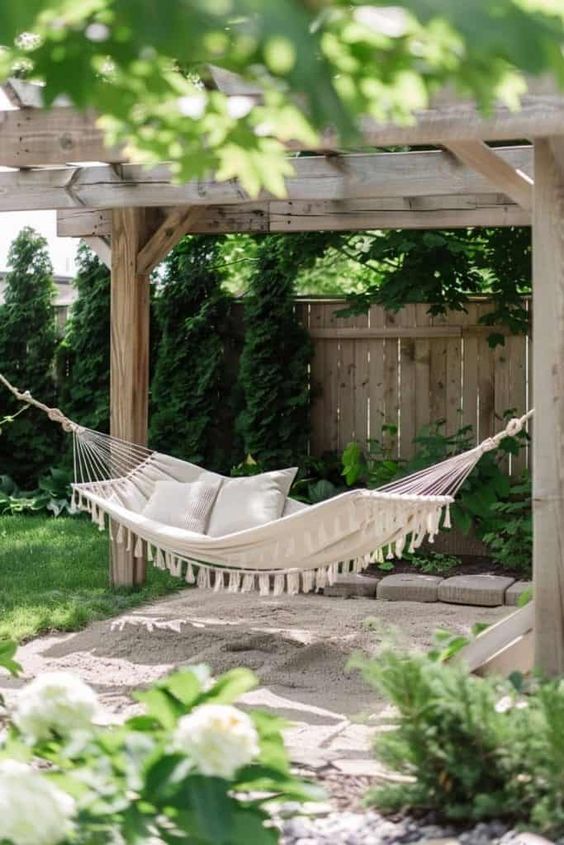 Cozy backyard hammock under wooden pergola with cushions, surrounded by lush greenery and flowers. Perfect outdoor relaxation spot.