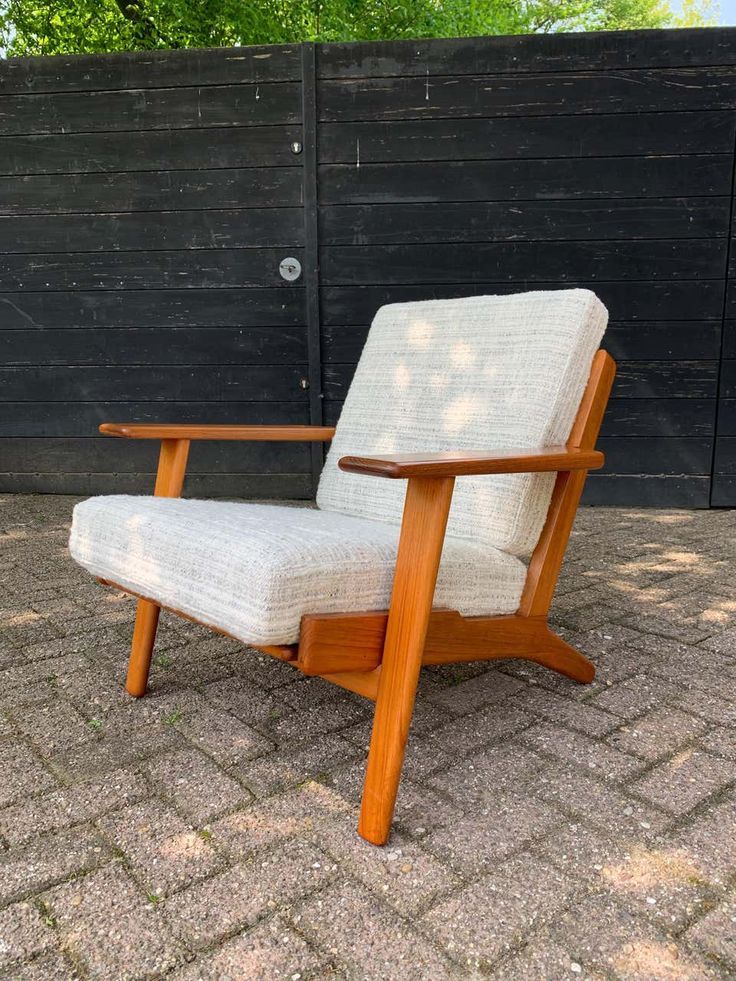 Mid-century modern wooden armchair with beige cushion set against a black wooden fence, outdoor setting.