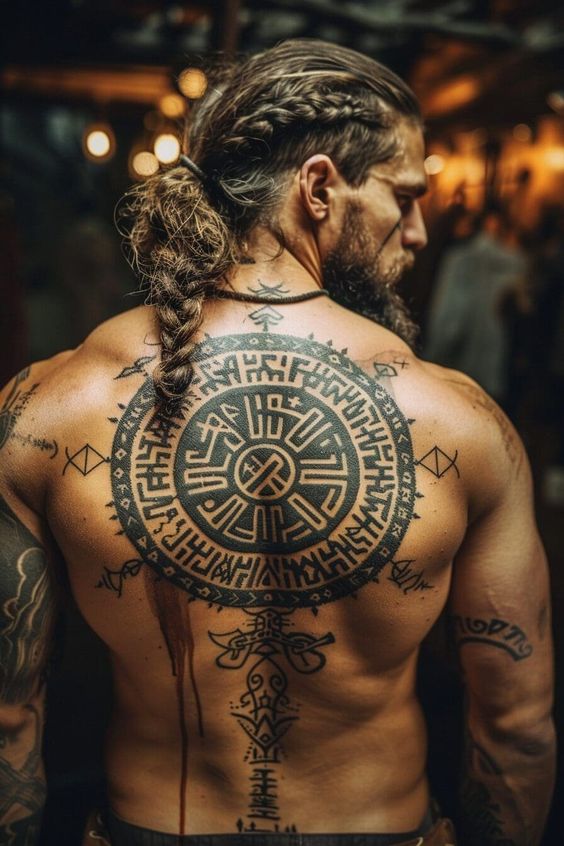 Man with intricate Viking-themed back tattoo and braided hair in dimly lit setting.