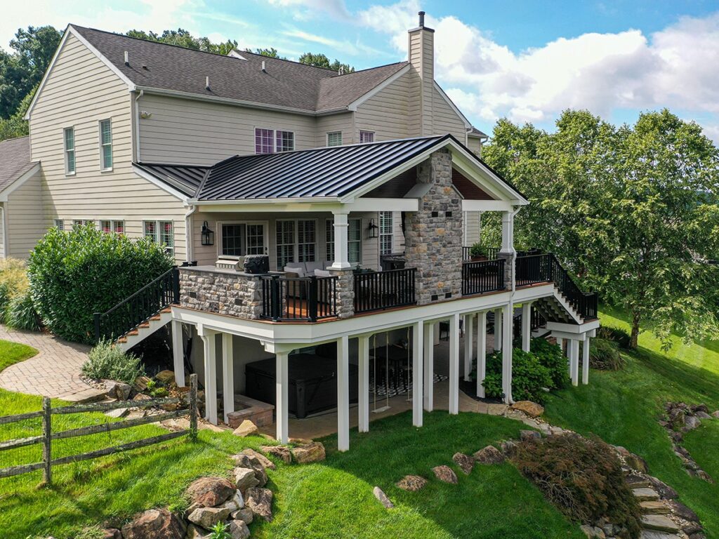 Spacious modern house with a stone chimney, elevated deck, and lush green garden on a sunny day.