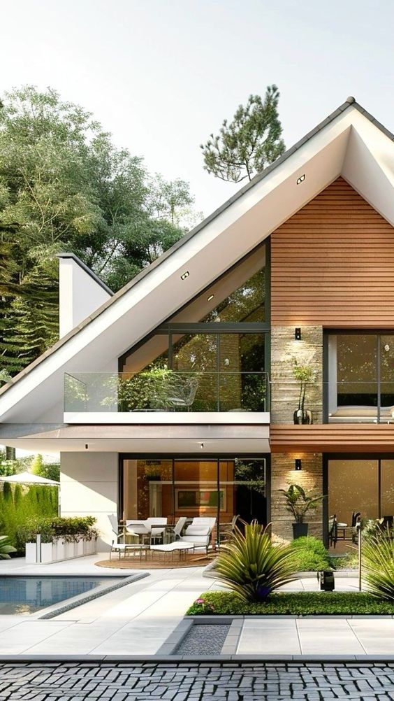 Modern A-frame house with large windows, poolside patio, and balcony surrounded by lush greenery.