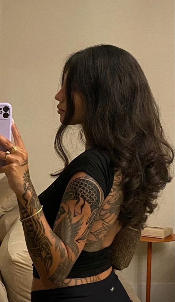 Side profile of a woman with long hair and tattoos, holding a purple smartphone in front of a mirror.