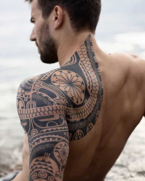 Man with intricate tribal tattoo covering his shoulder, arm, and back, sitting by the water's edge.