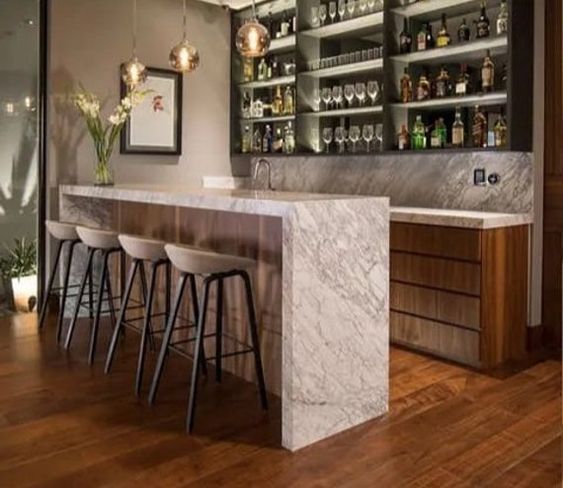 Contemporary home bar with marble countertop, bar stools, pendant lights, and shelves stocked with glasses and liquor bottles.