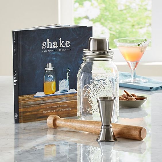 Cocktail book Shake, mason jar shaker, and cocktail tools on a countertop with a pink drink and almonds in background.