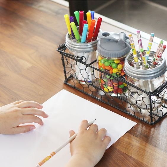 Child drawing with a pencil next to jars filled with colorful pens, pencils, and candy on a wooden table.