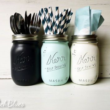 Three painted mason jars: black with utensils, mint with striped straws, and white with napkins, on a white wooden background.