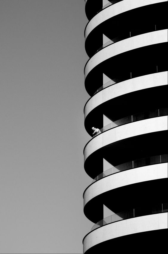 Black and white photo of modern round building with figure on balcony against clear sky, emphasizing urban architecture.