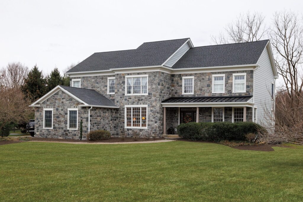 Elegant two-story stone house with large windows, wrap-around porch, manicured lawn, and surrounding trees on a cloudy day.