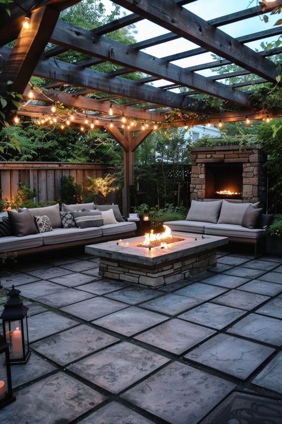 Cozy outdoor patio with fire pit, string lights, pergola, stone fireplace, and comfortable seating, ideal for evening relaxation.