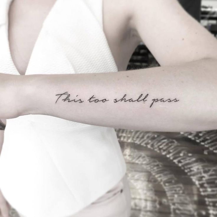 Forearm tattoo with cursive script that reads This too shall pass, shown on a person wearing a white sleeveless top.