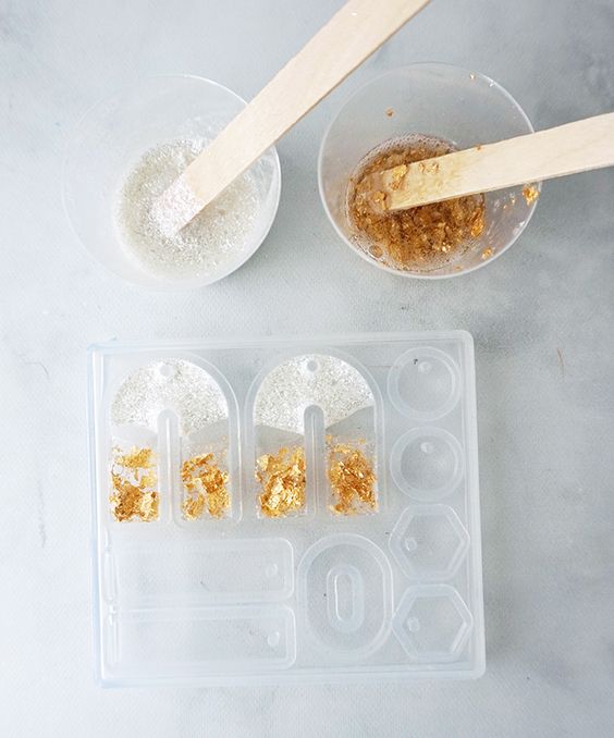 DIY resin crafting materials with mixing sticks, glitter, and gold foil arranged near a silicone mold on a white surface.