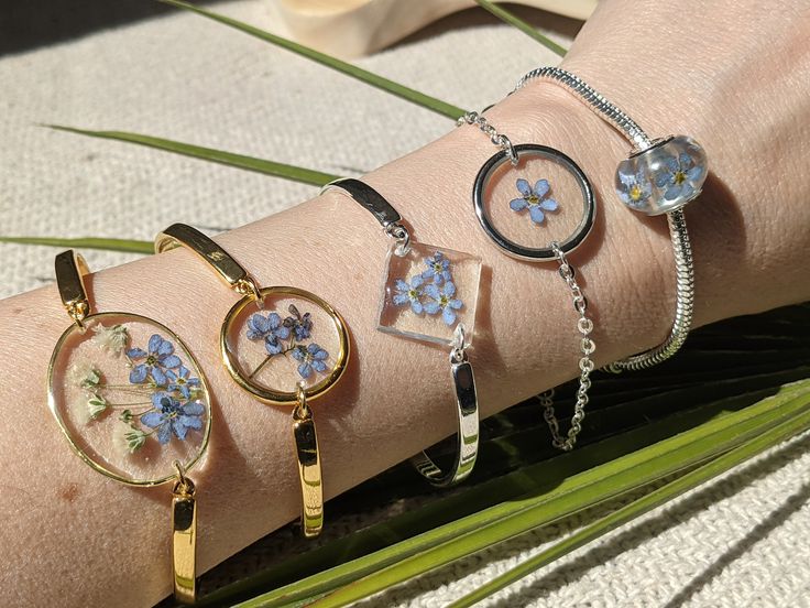 Hand wearing gold and silver resin bracelets with pressed blue flowers over a textured background, displaying floral jewelry.