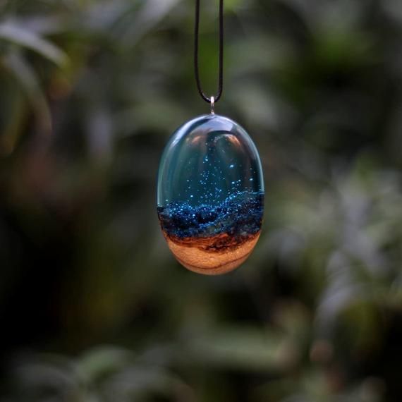 Handmade resin and wood pendant with a miniature ocean scene, capturing starry night sky, on a black cord against greenery.