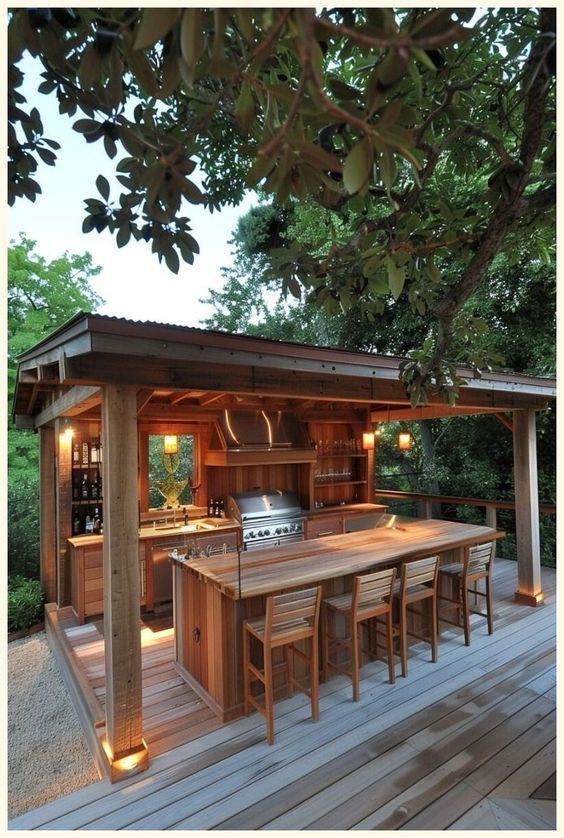 Outdoor wooden kitchen with bar seating, stainless steel appliances, and warm lighting under a canopy amidst lush greenery.