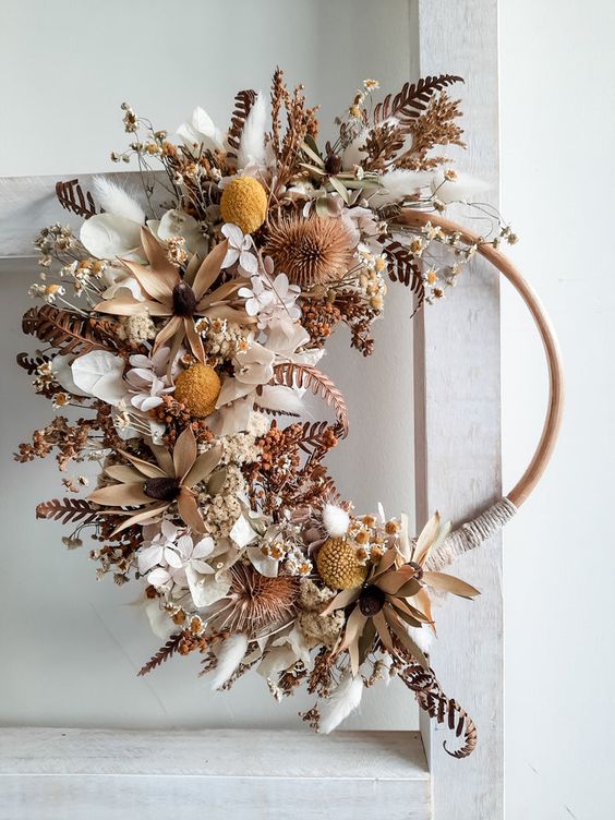 Rustic hoop wreath with dried flowers and foliage, featuring neutral shades and yellow accents, hung on a light background.