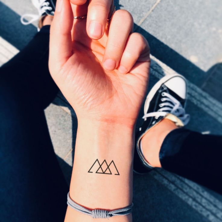 Minimalist geometric mountain tattoo on a wrist, with legs and black sneakers in the background.