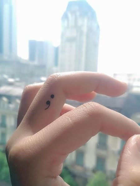 Close-up of a hand with a semicolon tattoo on the finger, against a blurred background of city buildings.