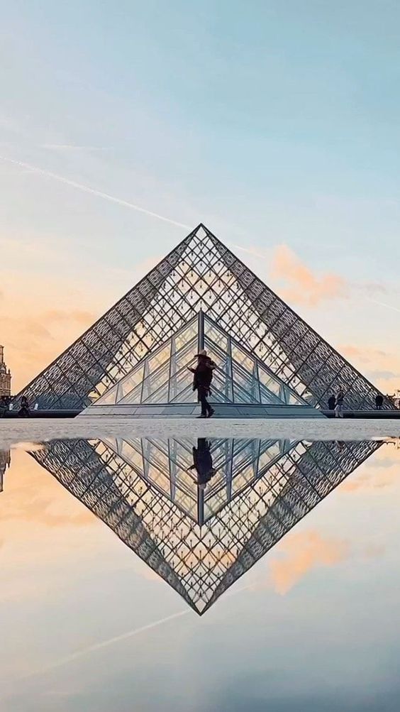 Glass pyramid of the Louvre Museum in Paris reflected in water at sunset with a person standing in front.