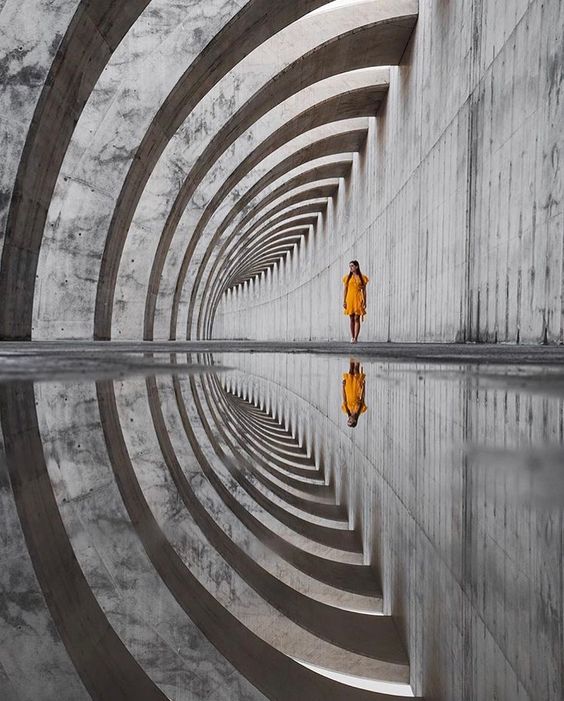 Woman in orange dress walking through concrete arches, reflection on water creating symmetrical pattern.