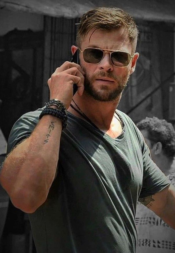 Man in green shirt with sunglasses, talking on phone, outside in casual setting, showing tattoos and bracelets on his left arm.