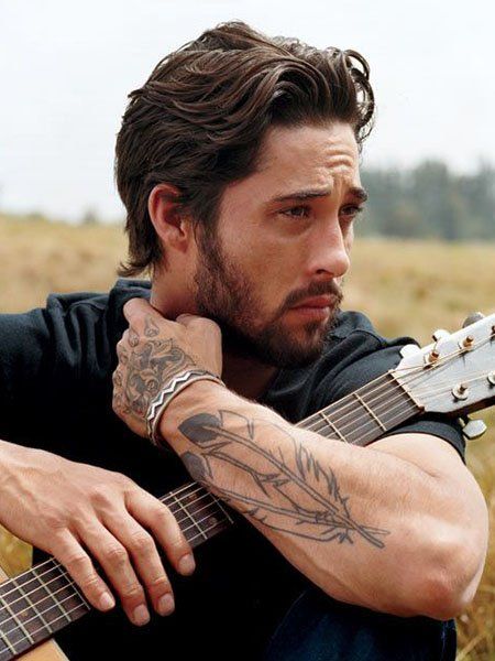 Man seated outdoors holding a guitar, with visible arm tattoos and pensive expression, background of fields and trees.