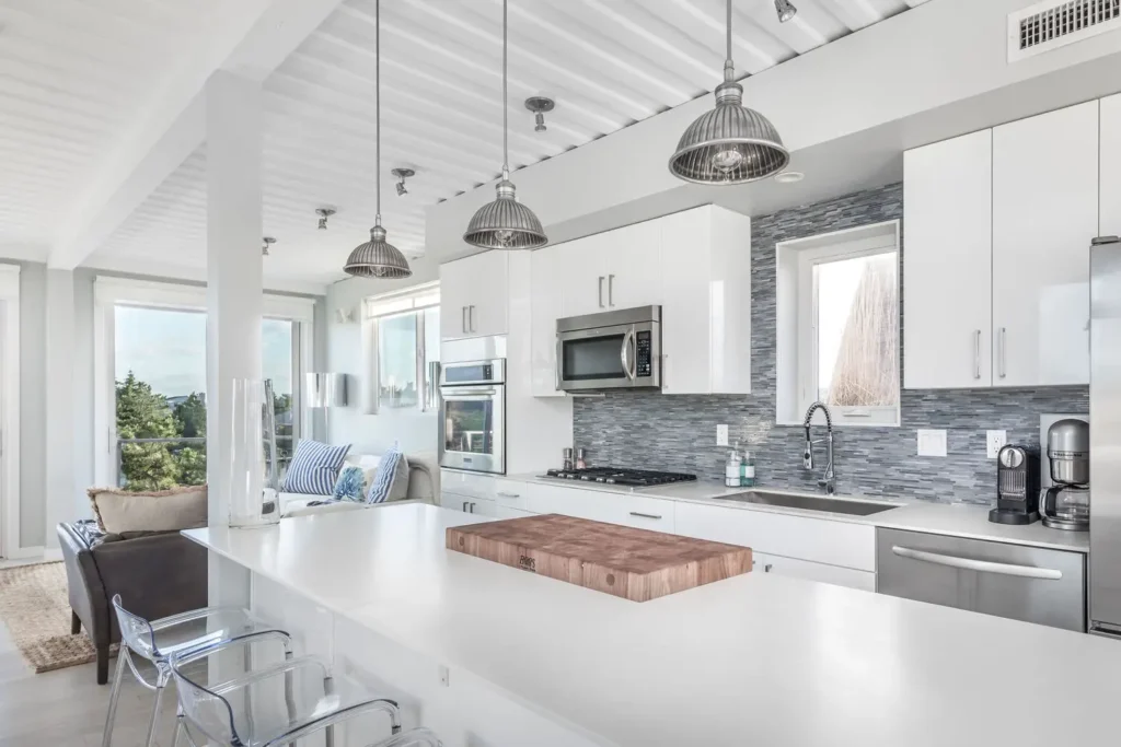 Modern kitchen with white cabinets, stainless steel appliances, large island, pendant lighting, and a cozy living area with windows.