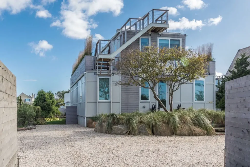 Modern beach house with rooftop deck, large windows, and landscaped yard under a blue sky with scattered clouds.