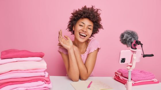 Woman vlogging with camera and microphone, surrounded by folded pink clothes, against pink background.