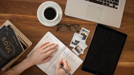 Person writing in journal at wooden desk with a laptop, coffee, glasses, books, and photos for creative thinking and planning.