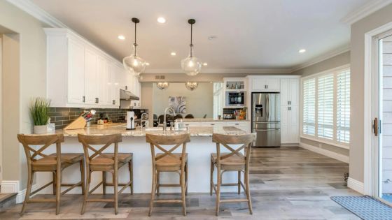 Modern kitchen with wooden bar stools, pendant lighting, white cabinets, stainless steel appliances, and hardwood floors.