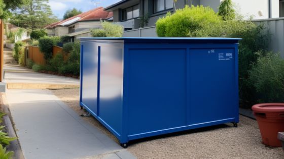 Blue metal storage container placed on a residential walkway, surrounded by greenery and houses in the background.