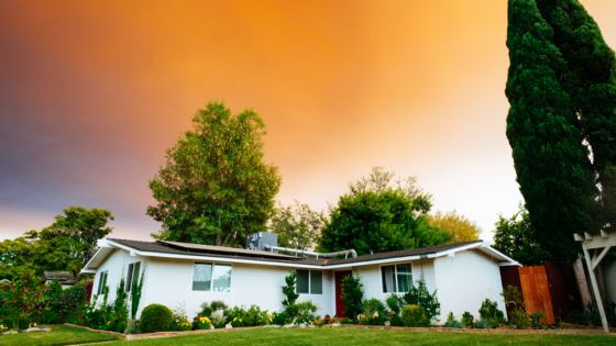 Charming white house with lush garden under vibrant orange sunset sky, surrounded by tall trees and greenery.