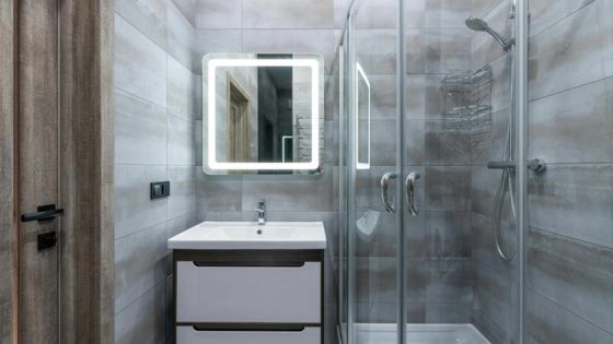 Modern bathroom with LED mirror, sleek sink cabinet, and glass-enclosed shower. Stylish gray tiled walls.